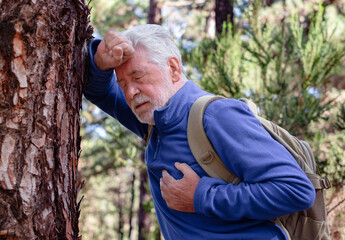 Senior man hiking in a forest pauses in distress leaning against a tree while holding his chest....
