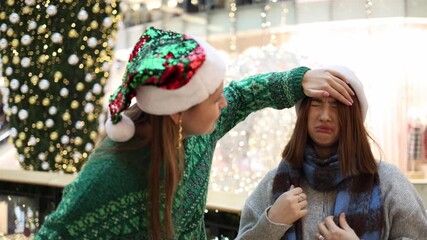 Two female friends in santa hats one checking forehead of another who feels unwell showing care support and friendship during cozy christmas holiday atmosphere
