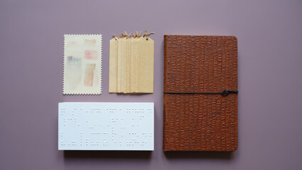 Artistic Flat Lay of Braille Slate, Textured Notebook, and Decorative Paper Elements on a Muted Purple Surface for Creative Projects and Visual Communication