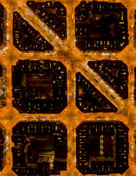 Aerial view of the grid-patterned streets illuminated in warm, golden light, contrasting with the dark building blocks of Eixample district, Barcelona, Catalonia, Spain.