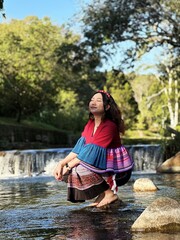 Joyful Asian woman in colorful Hmong fusion dress sitting by a scenic river.