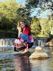 Joyful Asian woman in colorful Hmong fusion dress sitting by a scenic river.