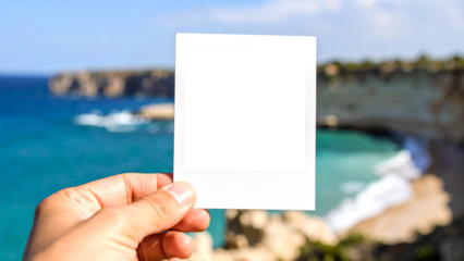 Hand holding a blank instant photo frame with a transparent background for easy overlay. The backdrop is a beautiful, blurred coastal scene with a turquoise sea, evoking travel memories.