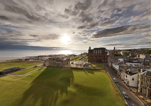Aerial view of golden sunlight kissing the lush green links contrasted against the rugged coastline and historic buildings of the town, St Andrews, Scotland, United Kingdom.