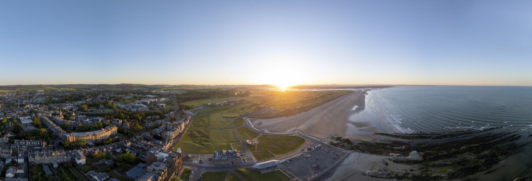 Aerial view of golden sunlight kissing the expansive West Sands beach and the historic golf course, contrasting with the cityscape, St Andrews, Scotland, United Kingdom.