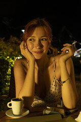 A high-quality lifestyle shot of an attractive young woman sitting at an outdoor cafe during a warm summer evening, enjoying a cup of coffee and smoking a cigarette. 