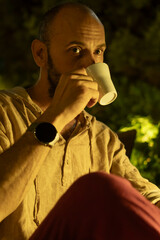 A portrait of a mature man sitting at a cozy outdoor cafe table during a warm summer evening, enjoying a cup of coffee. He looks relaxed and thoughtful in a modern urban lifestyle setting.