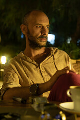 A mature man sitting at a cozy street cafe during a warm summer evening, enjoying a cup of coffee and a cigarette. He is portrayed in a relaxed, urban lifestyle setting with soft ambient lighting