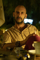 A mature man sitting at a cozy street cafe during a warm summer evening, enjoying a cup of coffee and a cigarette. He is portrayed in a relaxed, urban lifestyle setting with soft ambient lighting