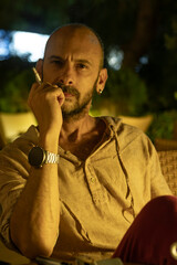 A mature man sitting at a cozy street cafe during a warm summer evening, enjoying a cup of coffee and a cigarette. He is portrayed in a relaxed, urban lifestyle setting with soft ambient lighting