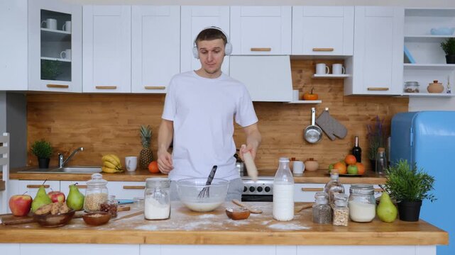 Happy young man wearing headphones, cooking and having fun dancing in a modern kitchen while preparing dough, playfully juggling a rolling pin and listening to his favorite music