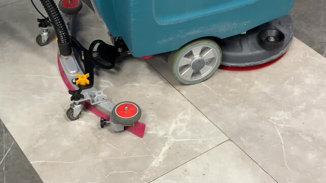 Industrial Floor Scrubber Cleaning Shiny Tiles Inside Corporate Lobby, Teal RideOn Machine With Red Rotary Brush, Wet Cleaning Path And Glossy Reflections, Maintenance Crew Oversight