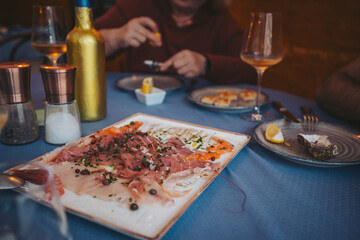 People are seated at a table in a restaurant, sharing a variety of food. One person is holding a lemon slice while another enjoys a drink. Plates contain different dishes to share