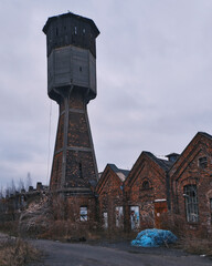 Old industrial water tower and abandoned brick factory buildings. A landscape of urban decay featuring weathered structures, historical masonry, and a gloomy overcast sky at a derelict site.