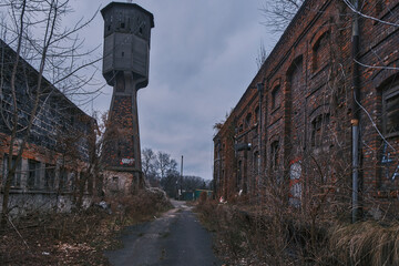 Old industrial water tower and abandoned brick factory buildings. A landscape of urban decay featuring weathered structures, historical masonry, and a gloomy overcast sky at a derelict site.