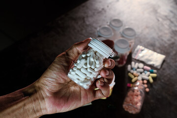 A person's hand holds a small prescription bottle overflowing with white oblong pills, symbolizing pharmaceutical reliance or the growing crisis of prescription drug abuse in a dimly lit setting.