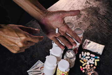Close-up of a hand presenting one white tablet amid various prescription pill containers, loose capsules, and cash.