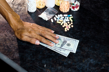 Hand reaching for or placing US currency near an assortment of pharmaceutical pills, capsules, and a small bag of white powder on a dark surface.