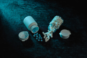 Various white capsules and blue tablets spill from two overturned pill bottles, symbolizing medication abuse, overdose risk, or pharmaceutical dependency under stark, dramatic blue lighting.