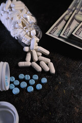 Assorted white and blue prescription tablets are scattered near an overturned clear pill bottle and several hundred-dollar bills on a dark surface.