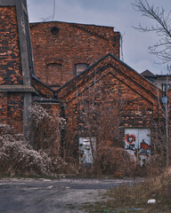 Abandoned industrial brick building with weathered walls and overgrown dry vegetation. Old factory ruins under a moody overcast sky, showcasing urban decay and historical architecture.