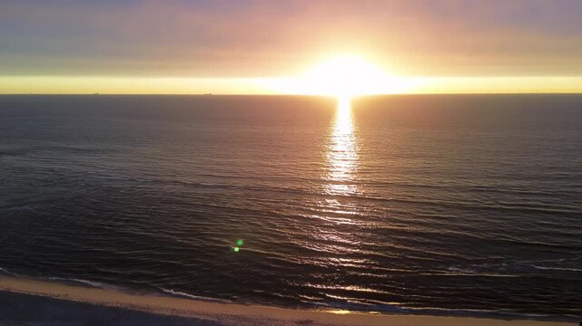 Aerial sunset beach view golden sun reflects on calm sea, silhouetted figures stroll shoreline under warm gradient sky, evoking serenity and depth.