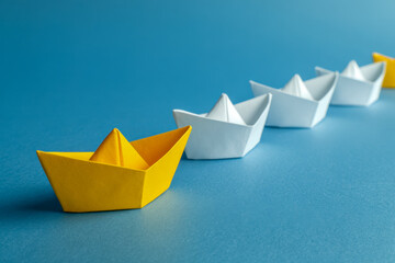 Yellow paper boat leading a line of white paper boats on blue surface symbolizing leadership and standing out in a crowd concept for business success