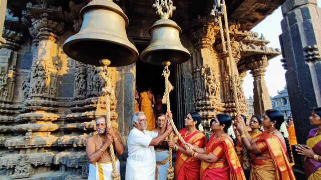 Worshippers are ringing large brass bells at an ornate hindu temple entrance, captured in a warm, spiritual low-angle composition.