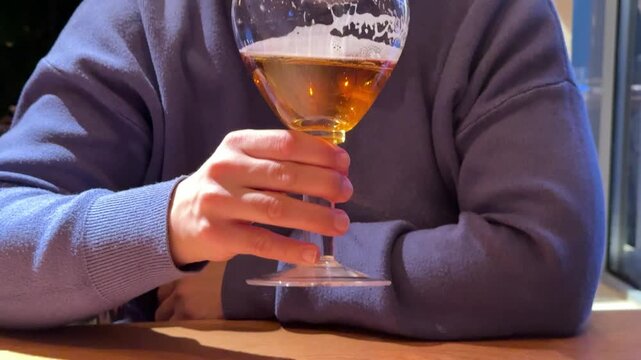 Hand Holding Wine Glass Amber Liquid, Phone On Wooden Table, Sweater Sleeve Visible, Warm Bistro Lighting And Soft Reflections On Glass. Freelancer Taking Quiet AfterWork Break, Checking