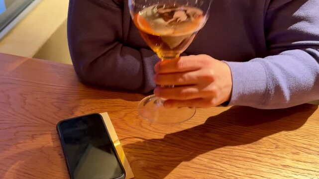 Hand Holding Wine Glass Amber Liquid, Phone On Wooden Table, Sweater Sleeve Visible, Warm Bistro Lighting And Soft Reflections On Glass. Freelancer Taking Quiet AfterWork Break, Checking
