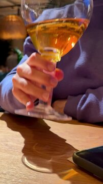 Hand Holding Wine Glass Amber Liquid, Phone On Wooden Table, Sweater Sleeve Visible, Warm Bistro Lighting And Soft Reflections On Glass. Freelancer Taking Quiet AfterWork Break, Checking