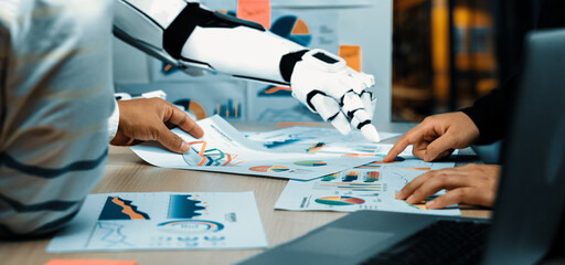 A human and a robotic arm collaborate in a modern office setting, analyzing data and charts, showcasing the blend of technology and teamwork in professional environments.Noogenesis