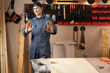 young carpenter say No thank you shows block, stop gesture while assemble furniture working in studio or workshop with tools on walls. woodworking workshop