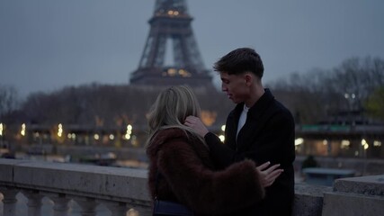 A young couple embraces intimately on a bridge in Paris France with the Eiffel Tower visible in the background at dusk The scene captures a romantic moment between the lovers in a popular travel desti