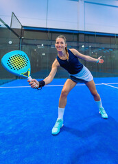 A beautiful, athletic female padel player leans in perfect position after a perfect wide shot with her racket, smiling beautifully on a blue indoor court.