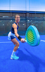 Beautiful young female padel player, wearing a skirt, posing with her legs bent in an athletic stance, happily and smilingly showing her blue racket, looking at the camera, on a blue indoor court.