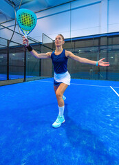 A confident and smiling female padel player maintains her balance with her arms outstretched after a perfect, winning shot on a blue indoor court.