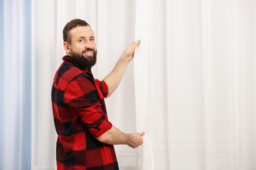 Man hanging up window curtains at home, space for text © New Africa