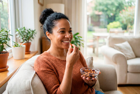 Smiling african american woman eating almonds while sitting comfortably on a sofa near a window