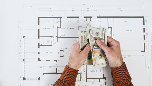 A man holds money over a paper house plan, wondering if he has enough money for a new home. The cost of home construction design services.