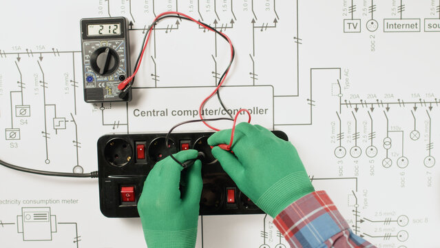 An electrician wearing gloves checks a large black extension cord stabilizer on a paper drawing of the electrical network for serviceability using a portable multimeter.
