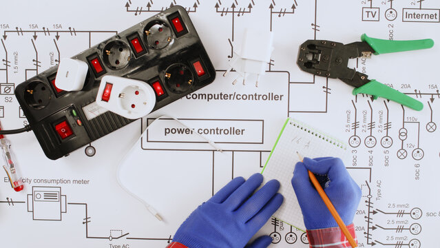 An electrical engineer writes down measurements on a large paper drawing of a house's electrical system, with a large uninterruptible power supply and network terminals lying nearby.