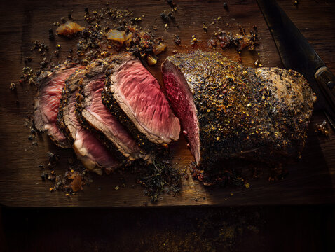 Top view of sliced roast beef on a rustic wooden cutting board. Juicy medium rare meat with peppercorn crust and knife. Holiday dinner concept