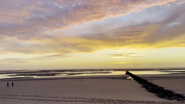 Sunset over Authie Bay in Berck, France, dramatic orange clouds above low tide sand flats, rocky groyne leading to the sea with walkers and dog silhouettes
