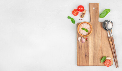 Minimal kitchen flat lay with wooden cutting board, tomatoes, basil, salt and utensils on white...
