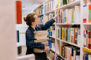woman is sitting at shelves in library, student is choosing book. library is filled with books. education, scientific work. university lecturer. young Caucasian woman is getting education.