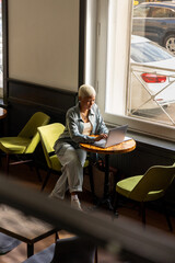 African American woman with white hair sits at desk with laptop, is surfing the Internet. female blogger, student, online education. lady works in coffee shop or coworking, top view.