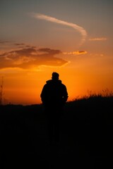 A solitary figure stands in silhouette against a breathtaking sunset, with warm golden and orange hues painting the dramatic sky above a dark horizon line. The vibrant evening light casts a powerful g