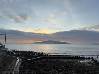 Fototapeta premium Sunrise Island Paradise: A distant island silhouette against a golden sunrise, with a rocky shore and calm ocean in the foreground under a blue sky with clouds.
