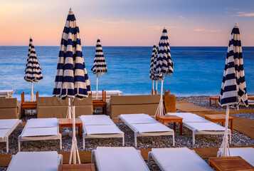 Blue and white striped umbrellas line a pebbled beach during a golden sunset over the Mediterranean...